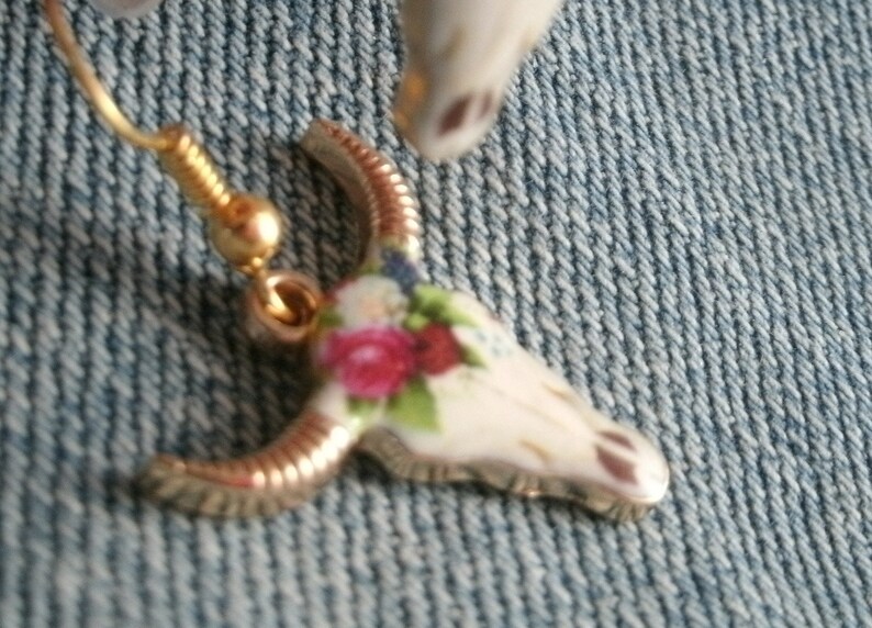 May include: Gold tone earrings with a white porcelain skull charm decorated with pink and blue floral details.