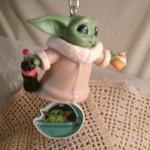 Op de afbeelding: Een groen en bruin beeldje van een personage uit de Star Wars-franchise, dat een flesje frisdrank en een klein taartje vasthoudt. Het beeldje draagt een roze jas met een witte bontkraag. Het beeldje is bevestigd aan een sleutelhanger met een klein beeldje van het personage in een ruimteschip.