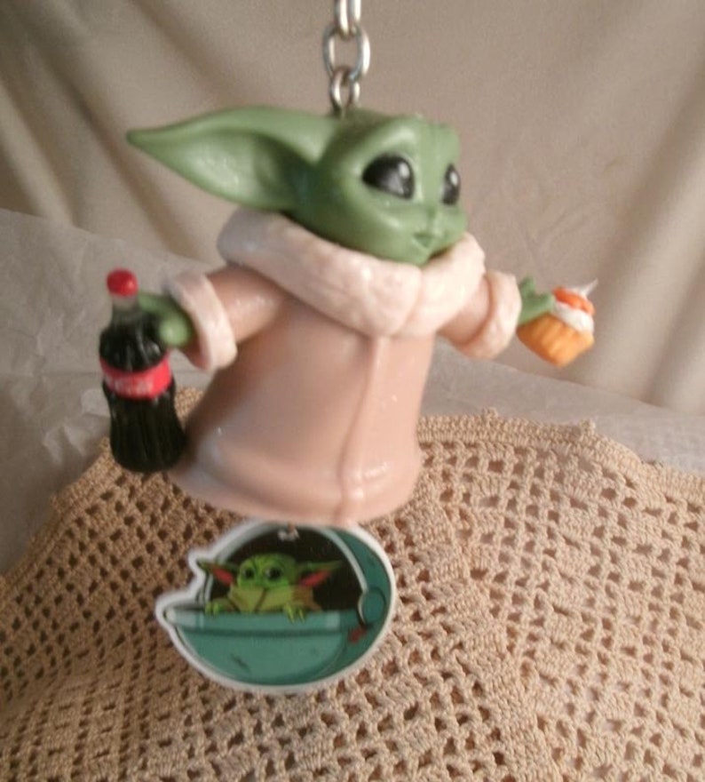Op de afbeelding: Een groen en bruin beeldje van een personage uit de Star Wars-franchise, dat een flesje frisdrank en een klein taartje vasthoudt. Het beeldje draagt een roze jas met een witte bontkraag. Het beeldje is bevestigd aan een sleutelhanger met een klein beeldje van het personage in een ruimteschip.