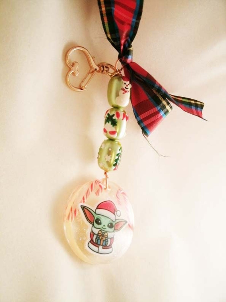 Peut inclure: Un porte-cl&eacute;s sur le th&egrave;me de No&euml;l avec un charme rond en r&eacute;sine transparente avec une image de dessin anim&eacute; d'un personnage vert portant un chapeau de P&egrave;re No&euml;l et tenant un cadeau. Le charme est attach&eacute; &agrave; un porte-cl&eacute;s dor&eacute; et une cha&icirc;ne de perles vertes et blanches avec des motifs sur le th&egrave;me de No&euml;l. Un ruban &eacute;cossais rouge et noir est nou&eacute; au sommet du porte-cl&eacute;s.