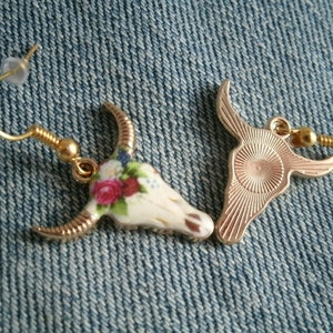 May include: A pair of gold tone earrings featuring a white ceramic bull skull design with floral accents. One earring has a solid gold tone back, while the other has a floral design.