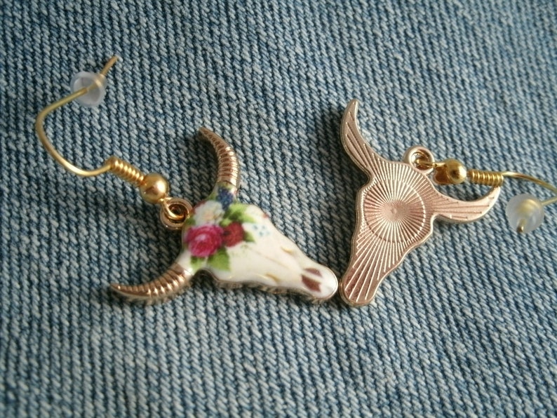 May include: A pair of gold tone earrings featuring a white ceramic bull skull design with floral accents. One earring has a solid gold tone back, while the other has a floral design.