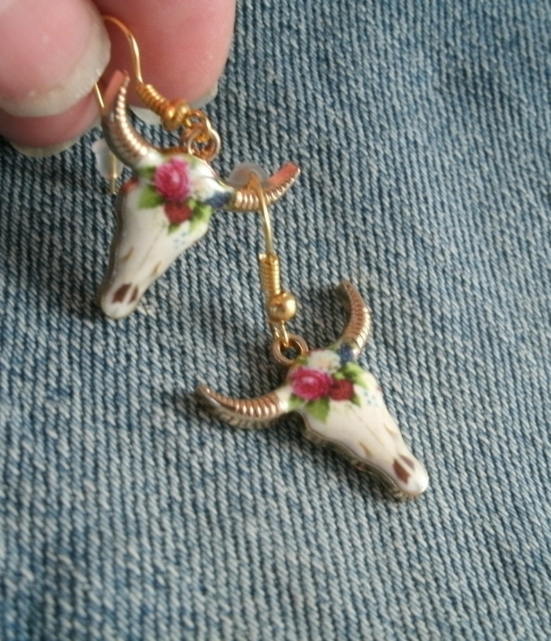 May include: A pair of gold tone earrings featuring a white ceramic bull skull design with floral accents. The earrings have a hook closure.