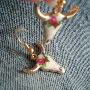 May include: A pair of gold tone earrings with a floral design. The earrings are shaped like a bull's head.