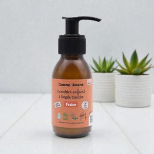 Op de afbeelding: Een bruine glazen fles met een zwarte pompdosering. Het etiket luidt "Comme Avant Dentifrice enfant à l'argile blanche Fraise". De fles staat op een wit oppervlak met twee kleine potplanten op de achtergrond.