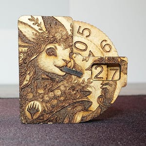 Può includere: Un contatore in legno con un'illustrazione di coniglio e un quadrante rotante con i numeri 5, 0, 6 e 27. Il contatore ha un logo di Magic: The Gathering.