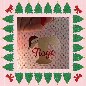 Peut inclure: Une décoration de Noël ronde et transparente avec le nom "Tiago" en script rouge, ornée d'un bonnet de Père Noël. L'ornement est suspendu par un ruban rouge. L'image est encadrée par des sapins verts et des nœuds rouges sur fond rose.