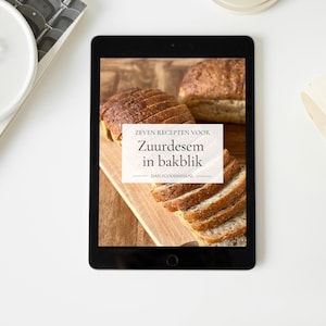 Puede incluir: Un iPad muestra una receta de pan de masa madre, con pan en rodajas sobre una tabla de madera. El texto en la pantalla dice "Zeven recepten voor Zuurdesem in bakblik". Un plato blanco y un recipiente están sobre la superficie blanca.
