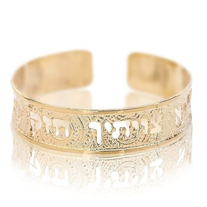 May include: Gold-toned cuff bracelet with Hebrew text and decorative patterns. The open cuff design features cut-out lettering and intricate detailing. The bracelet reflects light, creating a bright and elegant appearance.