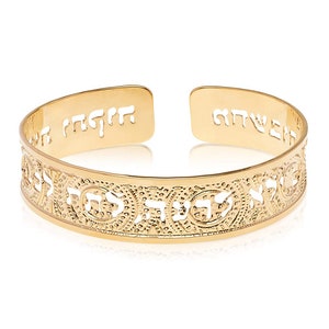 May include: A gold-colored cuff bracelet with Hebrew text. The open cuff design features intricate cut-out patterns and inscriptions. The bracelet is displayed on a white background, highlighting its polished finish and detailed craftsmanship.