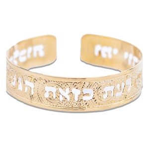 May include: Gold-toned cuff bracelet with white Hebrew lettering and decorative patterns. The open-ended bracelet features text along the outer band and on the inner edges. A piece of jewelry.