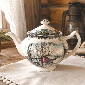 May include: A white ceramic teapot with a winter landscape scene. The teapot has a lid with floral detailing and a curved handle. The scene depicts bare trees, a red building, and a snowy landscape. The teapot is placed on a cream-coloured cloth.