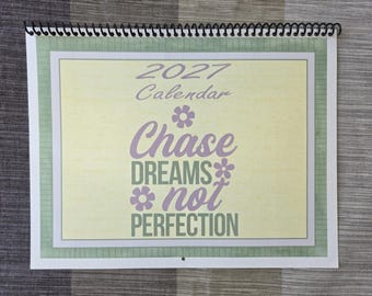 Pode incluir: Um calendário de parede para 2027 com encadernação em espiral no topo. A capa apresenta um fundo amarelo pálido com uma borda de grade verde. O texto diz 2027 Calendar e Chase DREAMS not PERFECTION em roxo e verde com ícones de flores.