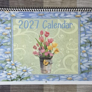 Peut inclure: Un calendrier 2027 avec une reliure spirale en haut. La couverture présente une illustration centrale d'un seau en métal rempli de tulipes roses, rouges et jaunes, agrémenté d'un papillon jaune. Le fond a un motif de volutes vert clair.
