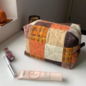 May include: A patchwork cosmetic bag in shades of orange, brown, and beige, with a zipper closure. The bag is next to a tube of Armani Skin Tint, a tube of mascara, and a lip gloss. The bag is on a white surface.