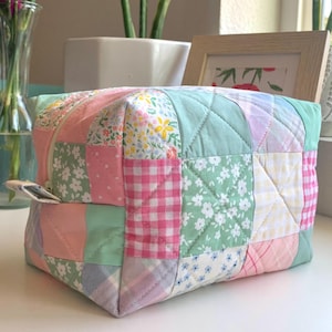 May include: A patchwork quilted cosmetic bag in pastel colors. The bag features a variety of fabrics, including floral prints, gingham, and solid colors, all sewn together in a square pattern. The bag has a zipper closure and a small fabric loop.
