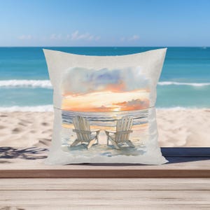 May include: A white throw pillow with a watercolor painting of two wooden chairs on a beach at sunset. The chairs are facing the ocean and the sky is a mix of orange, pink, and purple.