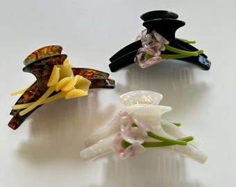 Könnte beinhalten: Drei Haarklammern mit Blumenakzenten. Eine Klammer ist schwarz, eine ist weiß und eine ist braun mit blauen und gelben Marmorierungen.