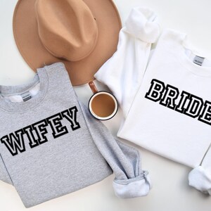 May include: A light gray sweatshirt with the word "WIFEY" in black block letters and a white sweatshirt with the word "BRIDE" in black block letters. A tan hat and a mug of coffee are also in the image.