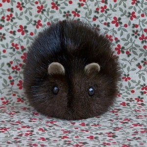 May include: A plush, round, dark brown hedgehog toy with small, light brown ears and black bead eyes. The soft toy is set against a floral patterned background with red flowers and green leaves.