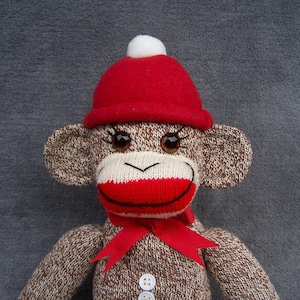 May include: A brown and white knit sock monkey doll featuring a red felt hat with a white pom-pom, a red ribbon tied around its neck, and two white buttons on its chest. The monkey has dark brown button eyes with black eyelashes.