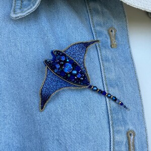 May include: A handcrafted, blue beaded stingray brooch. The brooch features a detailed design with various shades of blue beads and crystals, outlined with gold-colored thread. The brooch is attached to a light blue denim jacket.