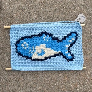 May include: A light blue rectangular crochet wall hanging featuring a dark blue and white fish graphic in the center. The piece is suspended between two horizontal wooden dowels and includes a small circular white tag with the text AN.
