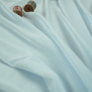 May include: A close-up shot of light blue fabric with three spools of thread. The fabric is draped, creating soft folds and shadows. The spools are brown and pink, adding a touch of color to the scene. The image has the text "lalathread".