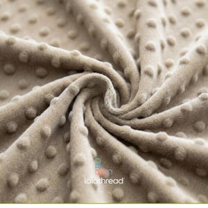 May include: A close-up shot of a beige fabric with a textured, dotted pattern. The fabric is folded and swirled, creating shadows and depth. The material appears plush and comfortable, suitable for blankets or clothing.
