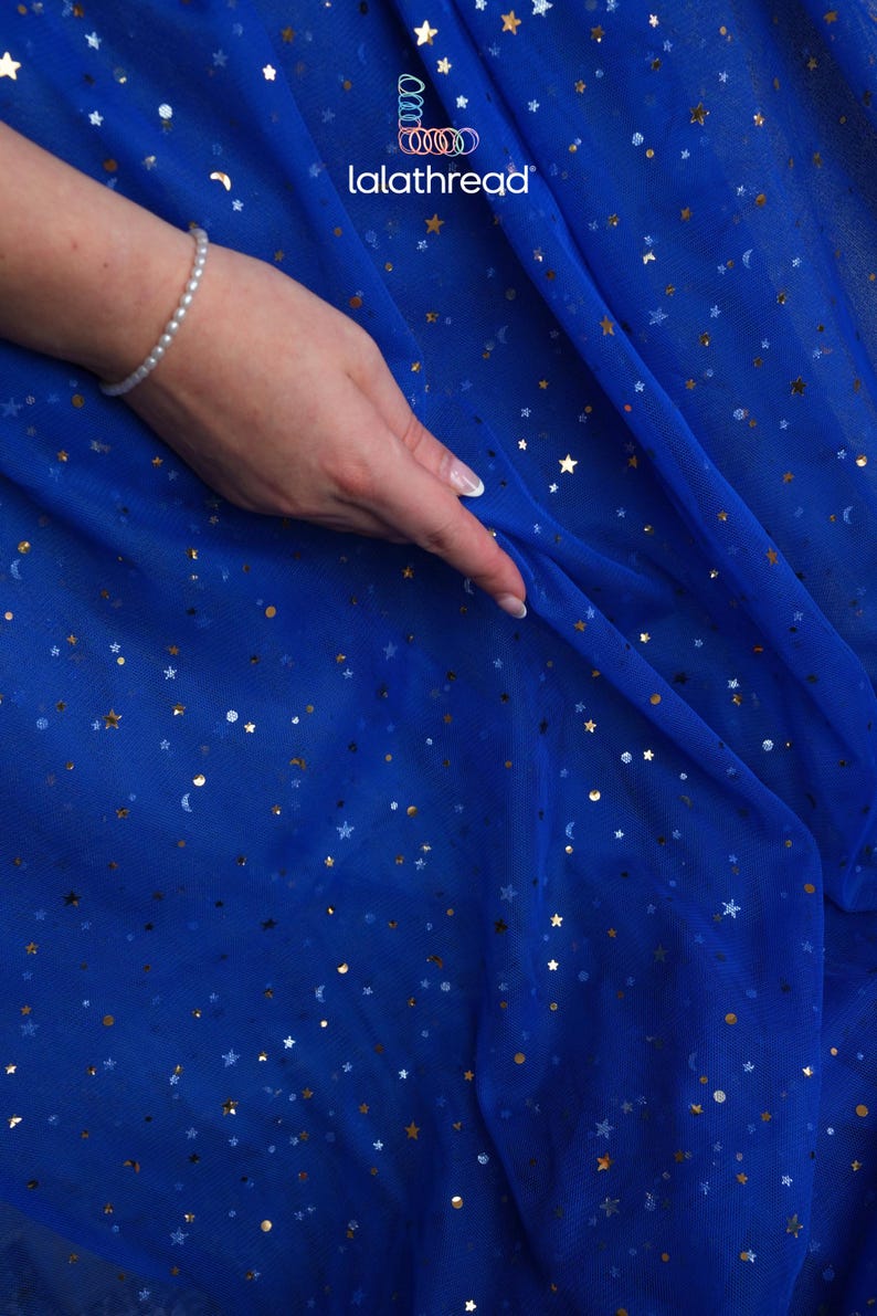May include: Royal blue fabric with gold stars and crescent moons. The fabric is draped, with a hand wearing a pearl bracelet touching the material. The logo "lalathread" is visible in the upper right corner.