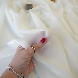 May include: A hand holding a swatch of sheer, off-white fabric. The fabric is draped in soft folds, with a slight sheen. The hand is adorned with a gold ring and a pearl bracelet. Two spools of thread are visible in the background.