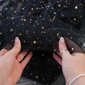 May include: Black tulle fabric with gold and silver star and moon sequins. The fabric is held up by two hands, showcasing the delicate, sheer material. The brand name "lalathread" is visible in the upper center.