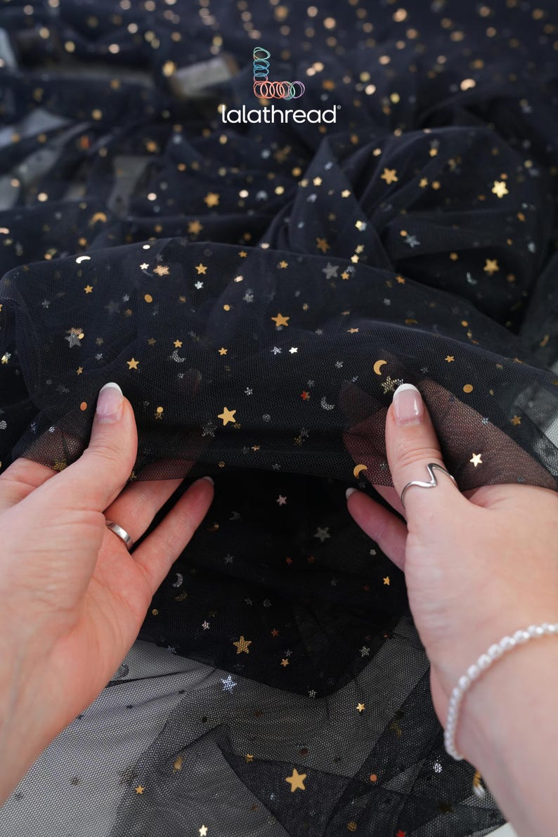 May include: Black tulle fabric with gold and silver star and moon sequins. The fabric is held up by two hands, showcasing the delicate, sheer material. The brand name "lalathread" is visible in the upper center.