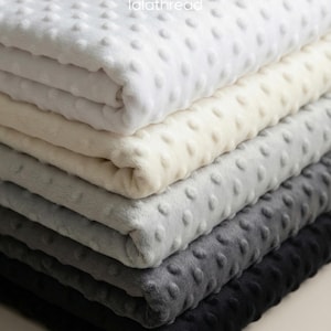 May include: A pile of soft blankets in shades of white, cream, grey, and black. Each blanket features a textured, dotted pattern. The blankets are neatly folded, displaying their soft and inviting texture.