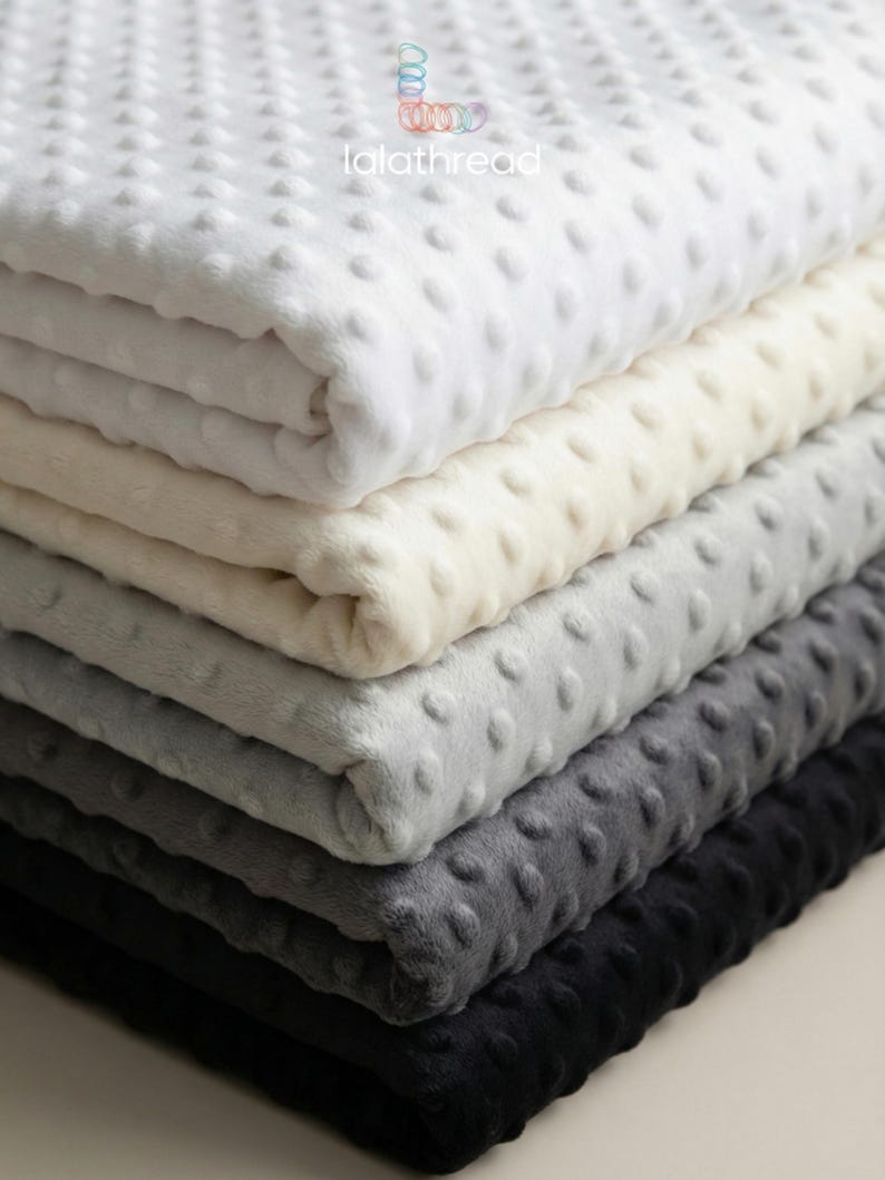 May include: A pile of soft blankets in shades of white, cream, grey, and black. Each blanket features a textured, dotted pattern. The blankets are neatly folded, displaying their soft and inviting texture.