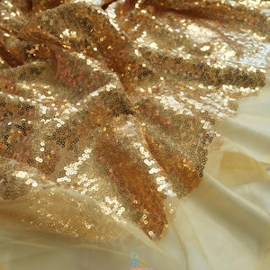 May include: Close-up of a gold sequin fabric with a soft, flowing texture. The sequins are densely packed, creating a shimmering effect. The fabric is layered over a lighter, sheer material. The logo "lalathread" is visible at the bottom.