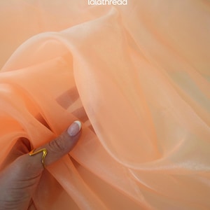 May include: Peach-colored sheer fabric with a soft, flowing texture. The fabric is semi-transparent, allowing light to pass through, and is gently held by a hand wearing a gold ring. The fabric has the text "lalathread" in the upper right corner.