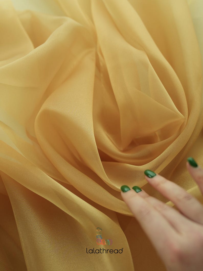 May include: A close-up of a sheer, golden yellow fabric. The fabric is draped and folded, creating soft, flowing lines. The fabric is translucent, allowing light to pass through it.