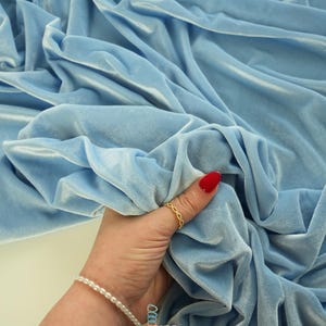 May include: A close-up of a light blue velvet fabric with soft folds. The fabric is being held by a hand wearing a gold ring and a pearl bracelet. The fabric has a shiny appearance. The logo "lalathread" is visible.
