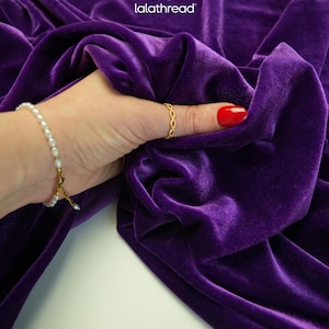 May include: Close-up of a luxurious, deep purple velvet fabric. The fabric is draped and held by a hand wearing a gold ring and a pearl bracelet. The brand name "lalathread" is visible in the upper right corner.