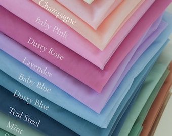 May include: A layered display of tulle fabric in various colors, including white, ivory, blush, champagne, baby pink, dusty rose, lavender, baby blue, dusty blue, teal steel, mint, sage green, caramel, and brown. The names of the colors are printed on the fabric.