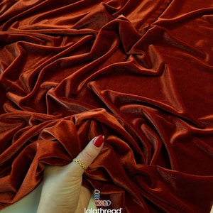 May include: Rust-colored stretch velvet fabric with visible folds and a soft sheen. The fabric is 58 inches wide and is accompanied by spools of thread. The text on the image reads "46 colors • stretch velvet fabric • 58" wide".