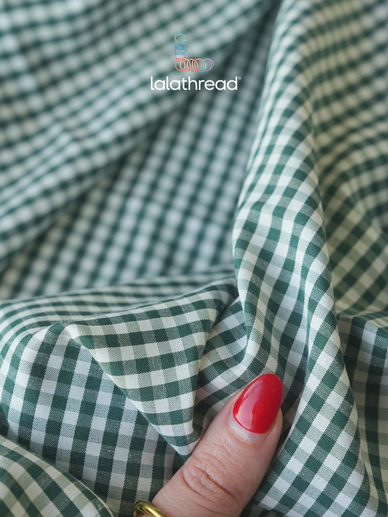 Puede incluir: Primer plano de tela de cuadros verdes y blancos. La tela est&aacute; doblada y drapeada, mostrando los detalles del patr&oacute;n. El nombre de la marca "lalathread" es visible. Una mano con esmalte de u&ntilde;as rojo toca la tela.