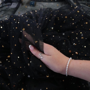 May include: Black tulle fabric with gold stars and crescent moons. The fabric is being held up, revealing its delicate texture. The brand name "lalathread" is visible in the upper left corner. A silver ring and a pearl bracelet are also visible.