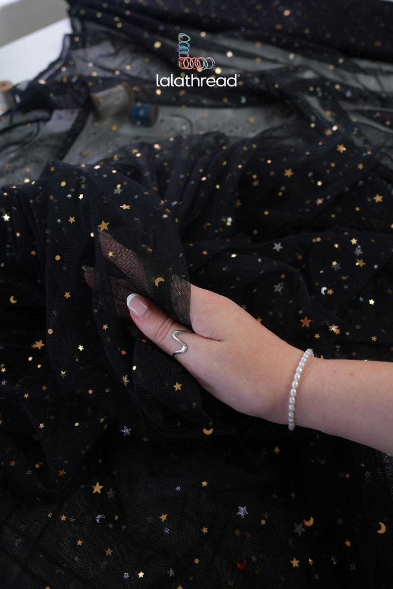 May include: Black tulle fabric with gold stars and crescent moons. The fabric is being held up, revealing its delicate texture. The brand name "lalathread" is visible in the upper left corner. A silver ring and a pearl bracelet are also visible.