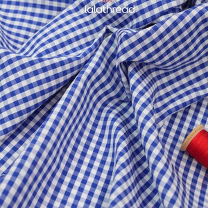 Puede incluir: Primer plano de tela de cuadros azul y blanco con el texto "lalathread" en blanco. La tela est&aacute; drapeada, mostrando pliegues y textura. Una bobina de hilo rojo es visible en la esquina inferior derecha.