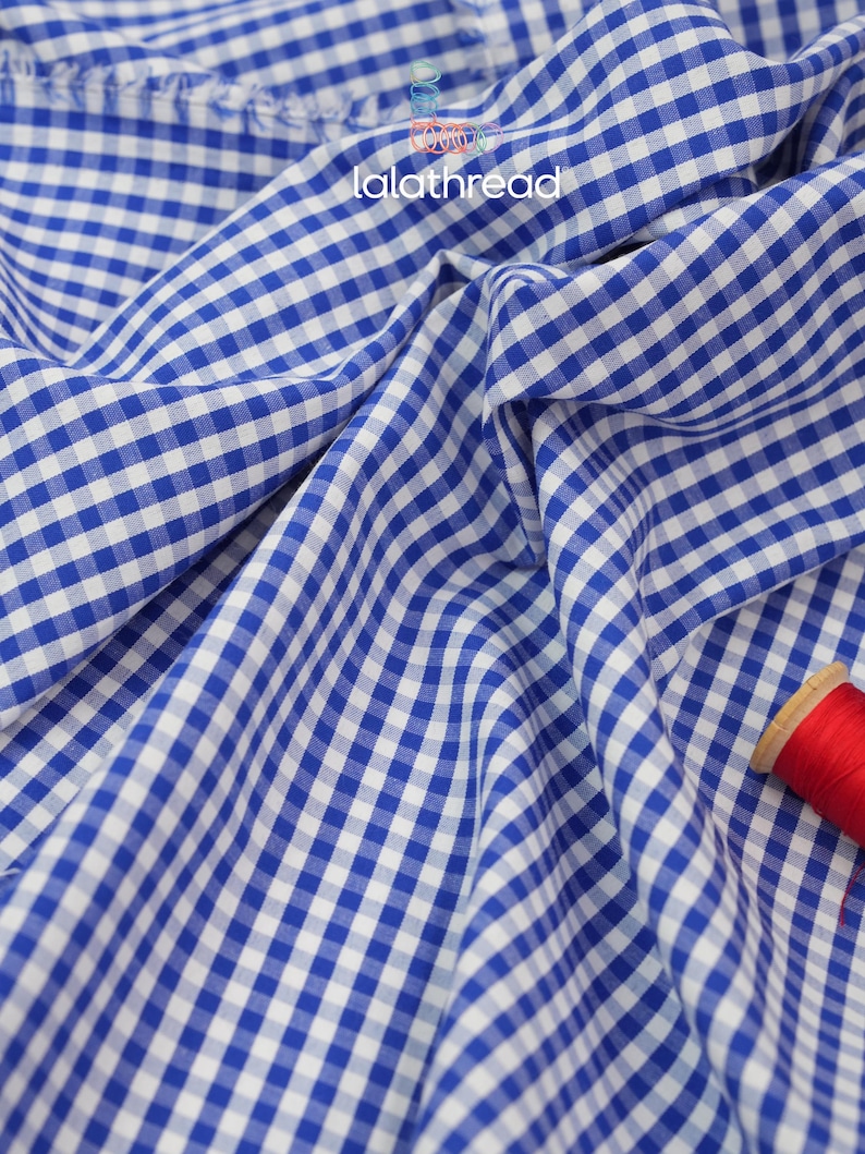 Puede incluir: Primer plano de tela de cuadros azul y blanco con el texto "lalathread" en blanco. La tela est&aacute; drapeada, mostrando pliegues y textura. Una bobina de hilo rojo es visible en la esquina inferior derecha.