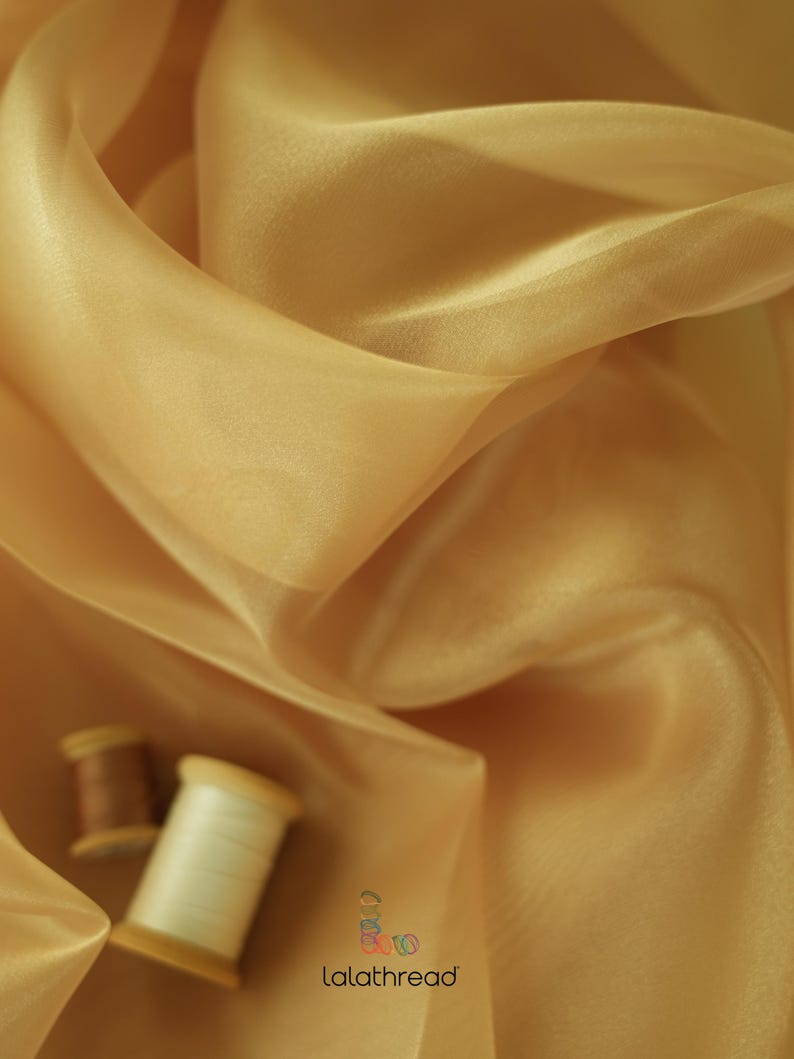 May include: A close-up of a golden sheer fabric, draped and folded. Two spools of thread are visible in the bottom left corner of the image. The text "lalathread" is visible in the bottom right corner of the image.