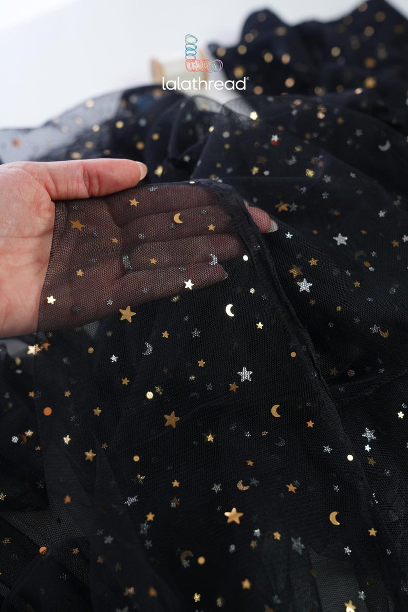 May include: Black tulle fabric with gold and silver star and crescent moon embellishments. The fabric is semi-sheer, with the brand name "lalathread" visible in the background. This fabric is suitable for apparel and craft projects.