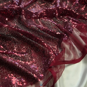 May include: Close-up of a burgundy sequin fabric. The fabric is covered in small, round sequins that catch the light, creating a shimmering effect. The fabric has a tulle backing and is likely used for apparel or decorative purposes.